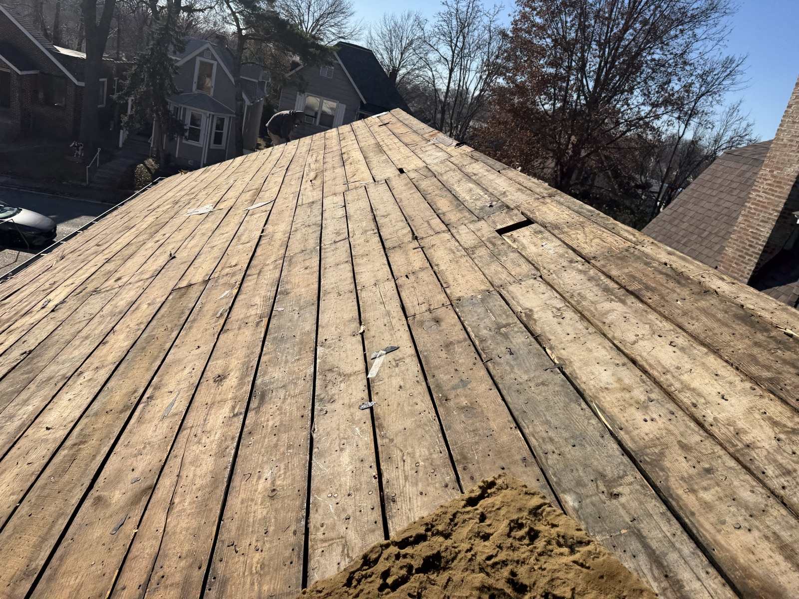 A roof stripped down to bare board decking, wide sawn planks running up to the ridge with gaps between them and one board split mid slope, with neighbouring houses and bare winter trees beyond