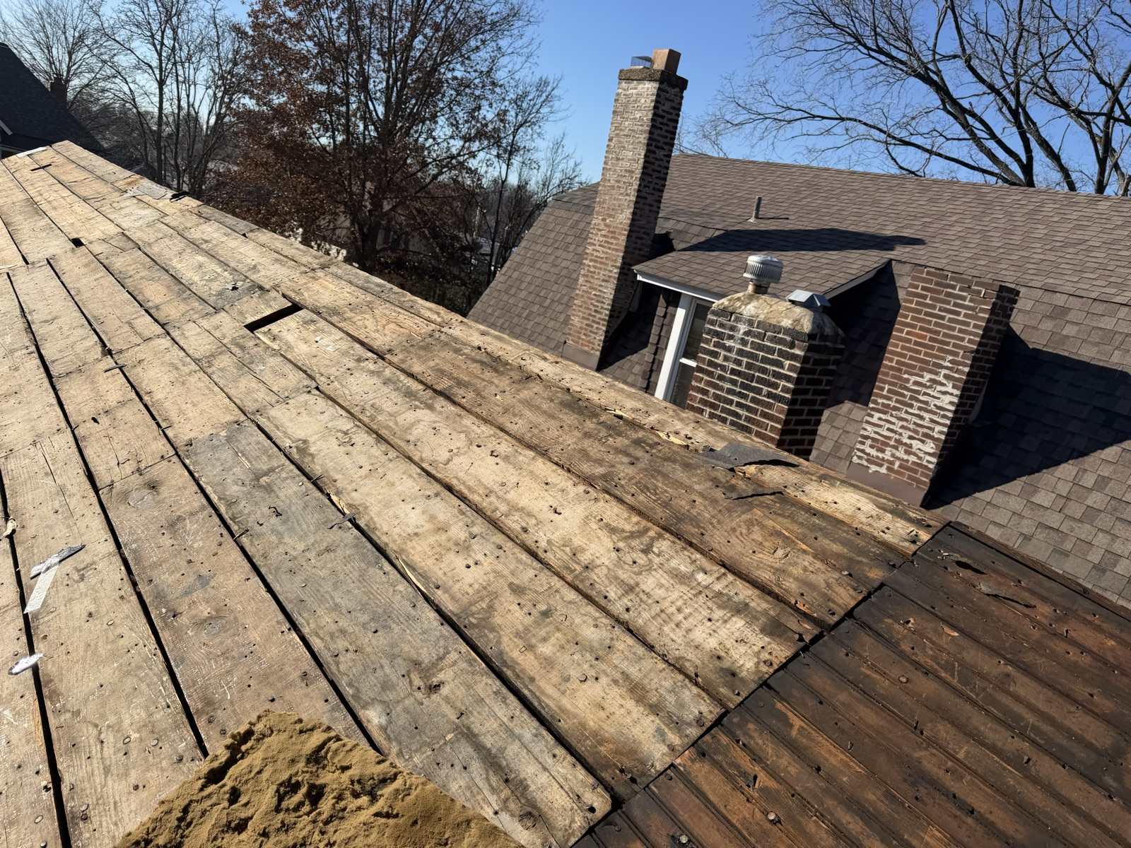A stripped roof plane of weathered wood planks in the foreground, looking across to the neighbouring brick house with two brick chimneys, one showing spalled and patched brickwork, under a clear winter sky