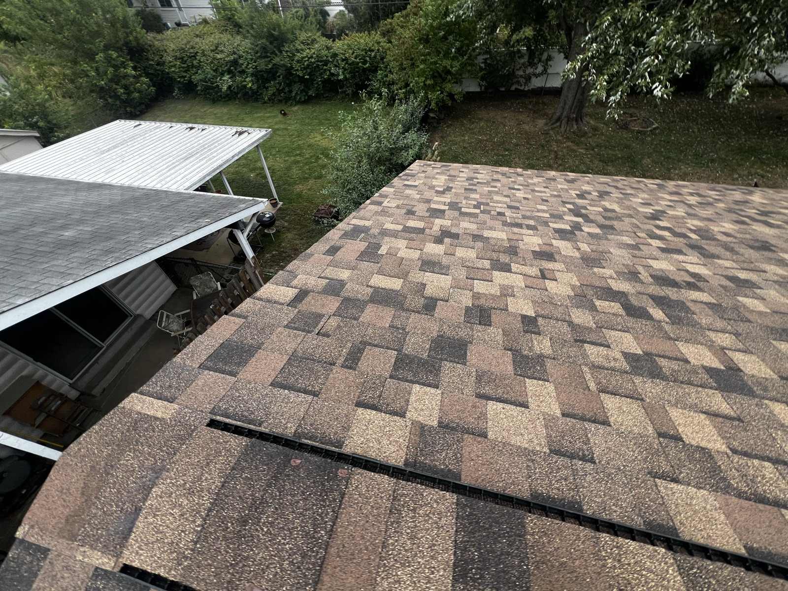 A completed multi-tone brown and tan architectural shingle roof seen from above, with a strip of ridge vent along the lower edge, an adjoining grey shingled porch roof and a white metal carport roof beside it, over a green back garden