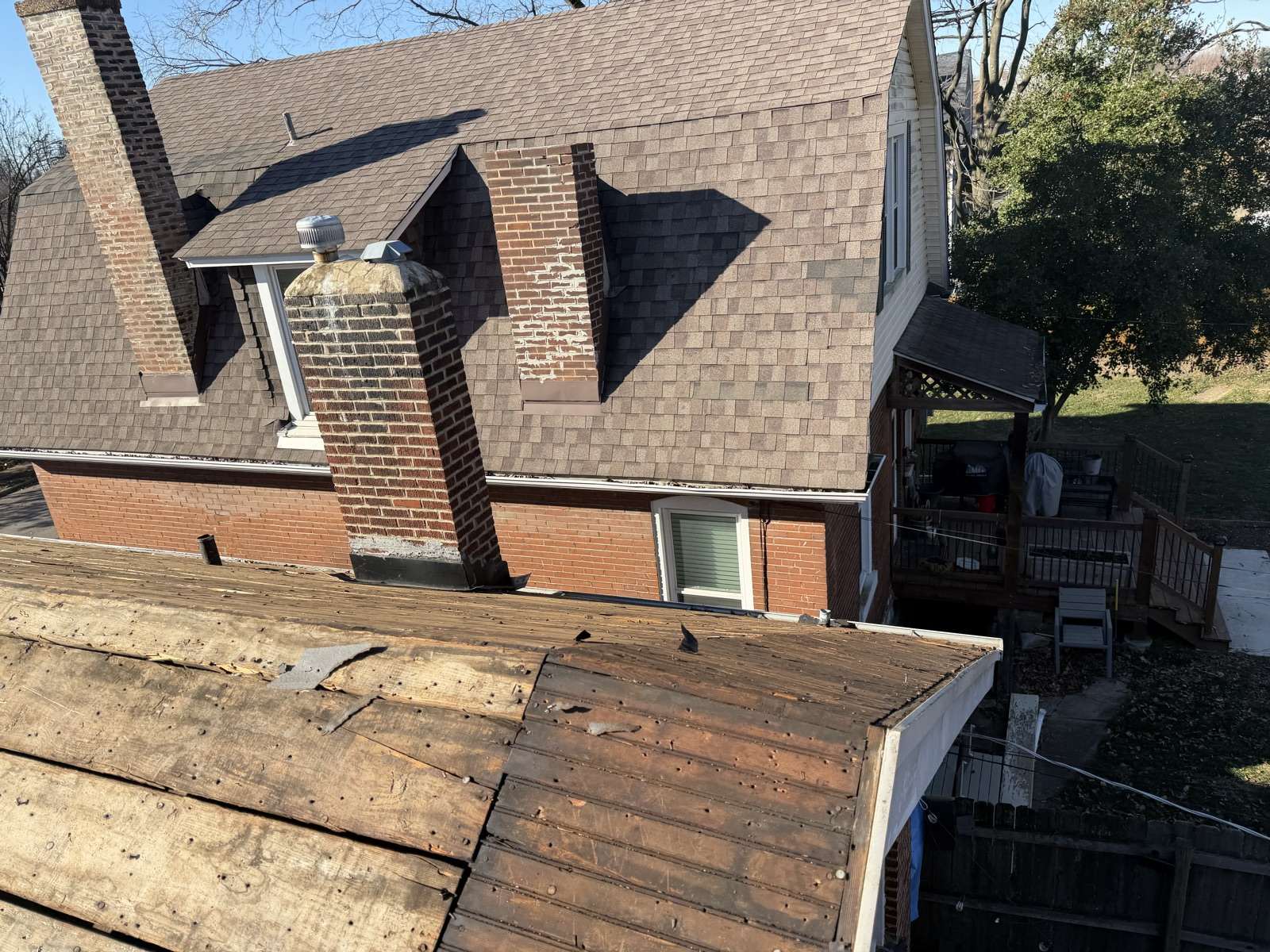 A lower porch roof stripped back to bare wood planks in the foreground, with the main brick house behind still carrying its old brown shingles, a spalling brick chimney and a wooden deck below