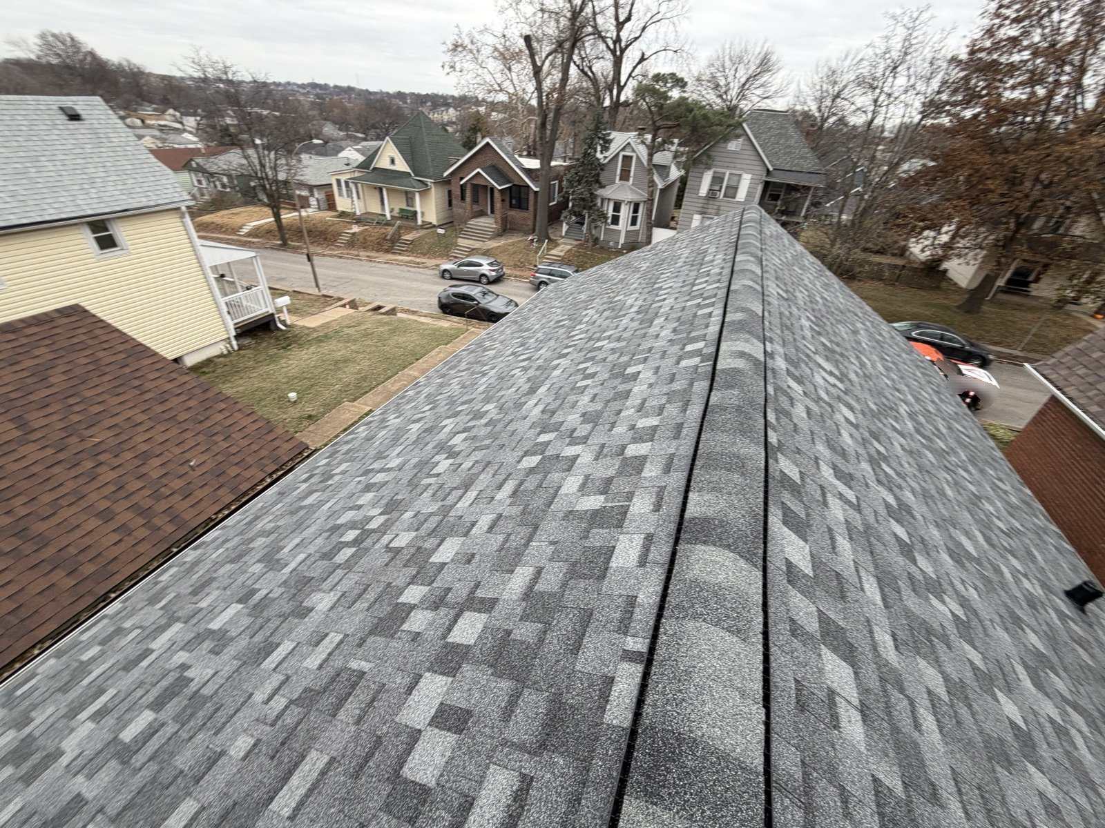Looking down the ridge of a newly shingled grey roof, with a continuous ridge vent along the peak and a street of older brick and frame St. Louis houses below on a bare winter day