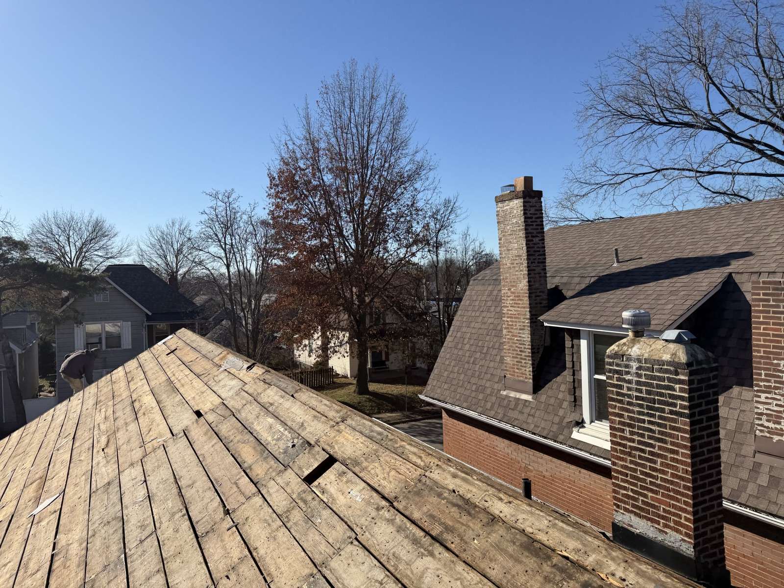 Standing on a stripped wood plank roof looking across to a neighbouring brick and shingle house with two chimneys and a metal roof vent, bare winter trees and a clear blue sky