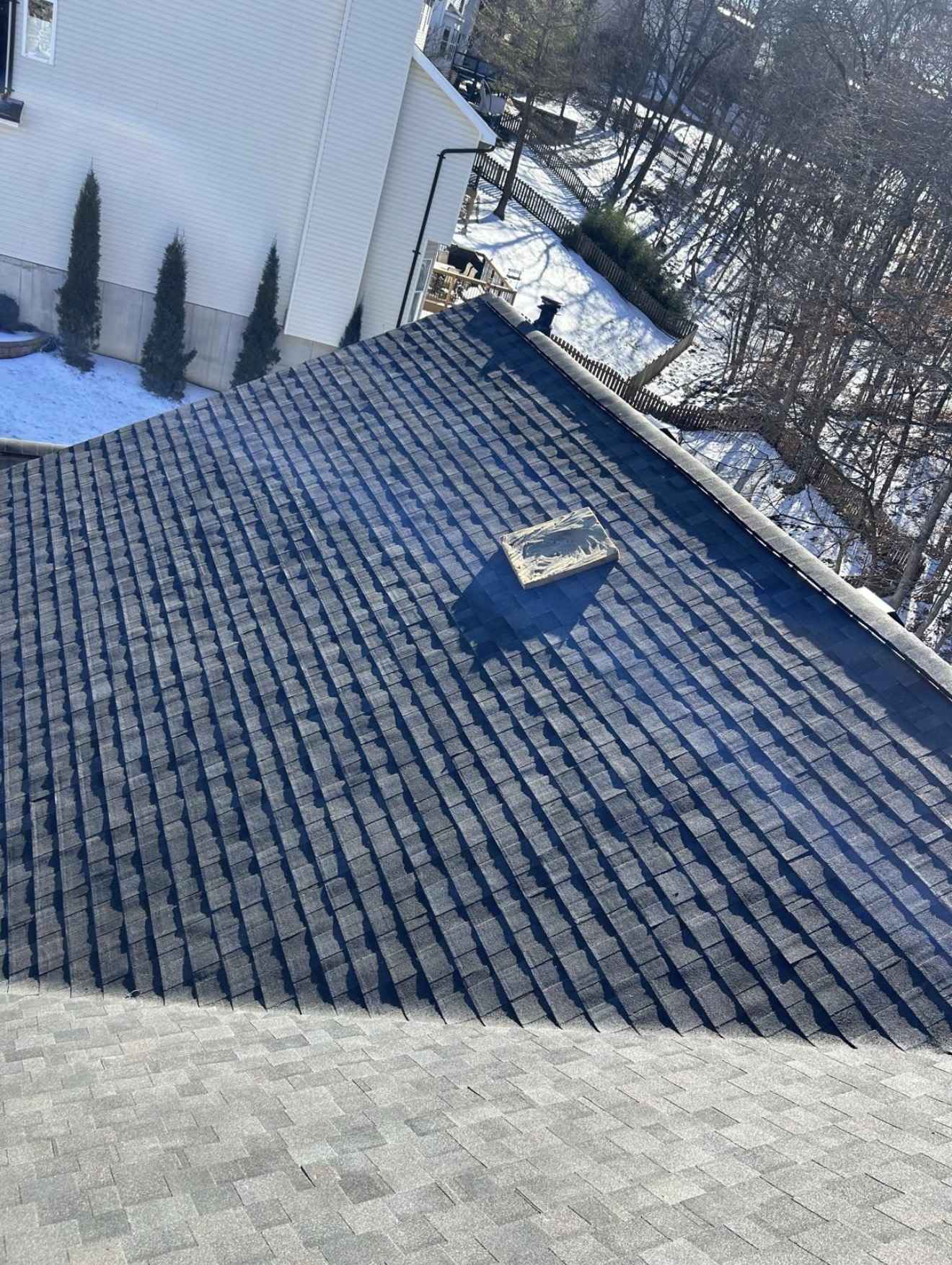 Dark laminate shingles being laid in staggered courses up a steep roof slope in winter, an unopened bundle resting mid slope, with a white sided house and snow covered ground behind