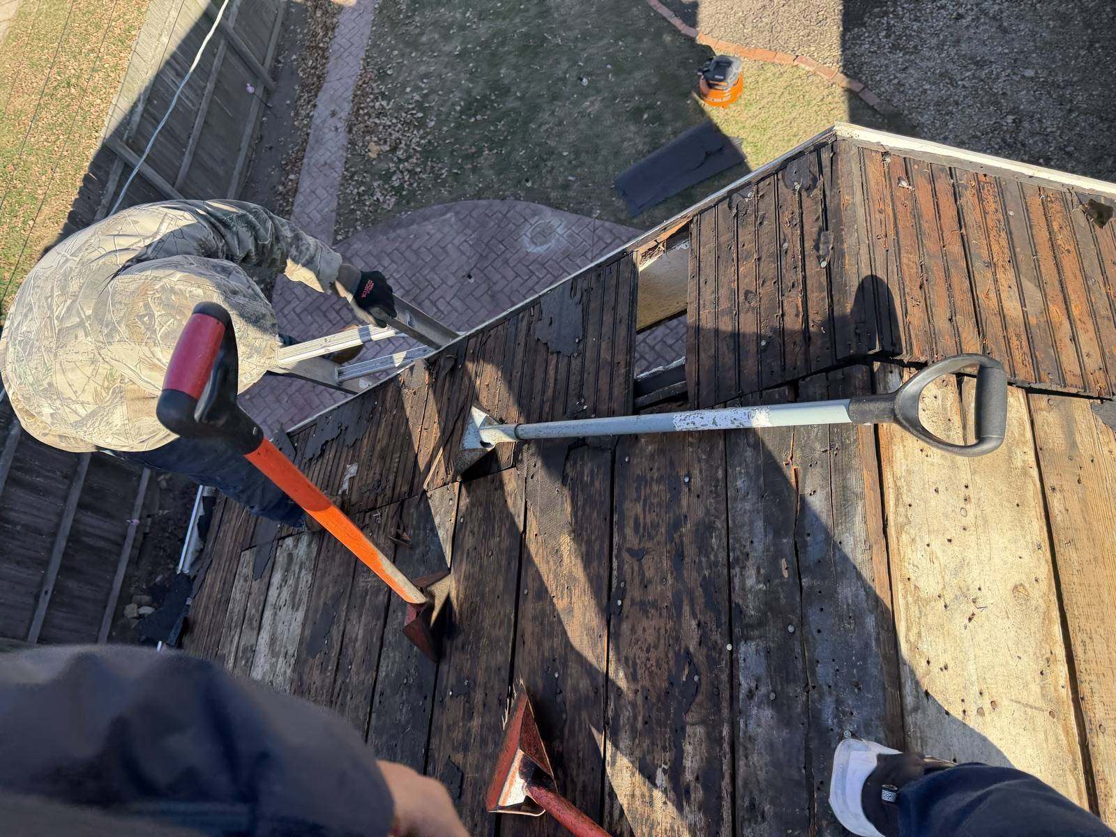 Looking straight down at a low roof being stripped to bare boards, two long handled tear-off spades and a pry bar lying on the deck, a ladder hooked over the eave and a brick patio below